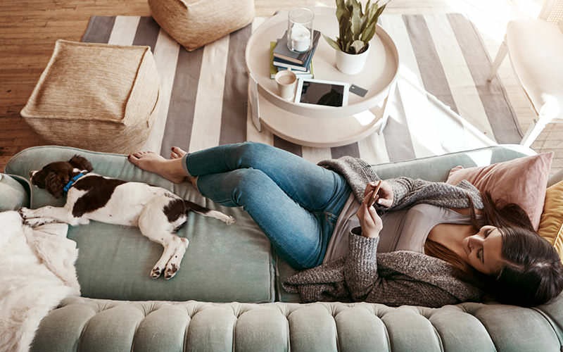 woman texts on her phone as she lies on her couch with her dog