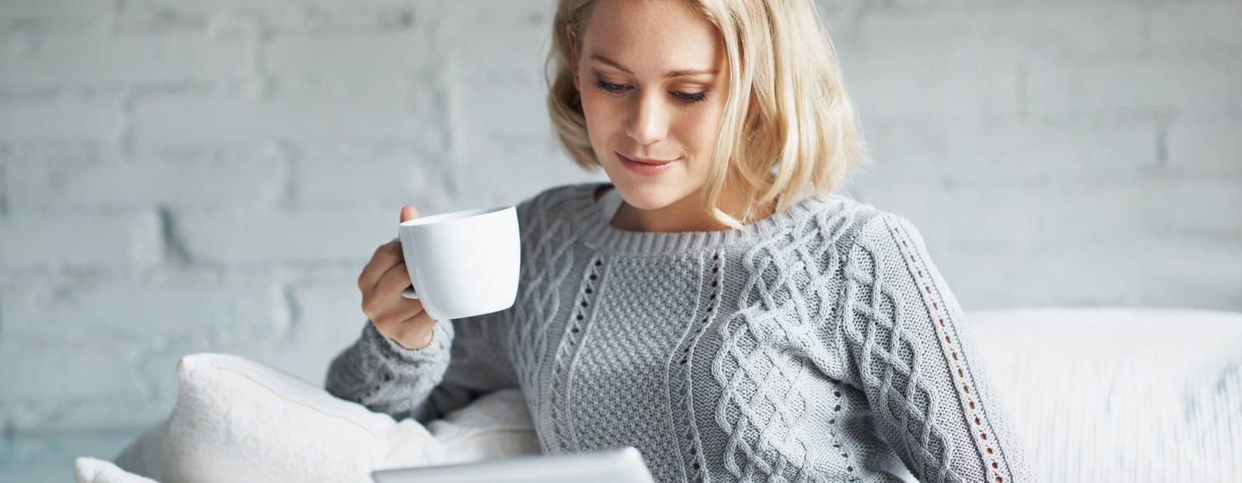 woman sits on her couch with a cup of coffee and looks at her tablet